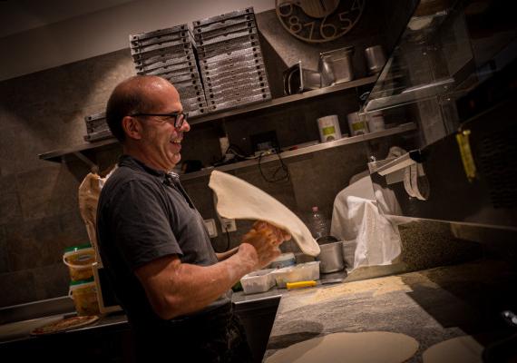 Un uomo con gli occhiali è in piedi davanti alla pizzeria SchartnerAlm e sorride amichevolmente alla telecamera